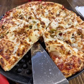 Pizza 9 - Veggie pizza - A cheese pizza with green peppers is placed on a black, textured tray on a wooden table. The pizza has a golden-brown crust and melted cheese. A metal pizza cutter is partially visible beneath the pizza. In the background, there is a red drink in a glass and a white napkin on the table. The setting appears to be a casual dining restaurant. - Pizza Near Me - Grants, New Mexico