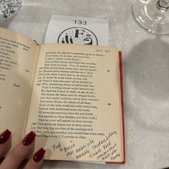 Flyleaf - Book that customers can write in when cashing out - The image shows a person with red painted nails holding an open book with printed text and handwritten notes on a page. The notes mention finding a person and a date. The table has a water bottle, a wine glass, and a paper with the number 133. The setting appears to be a cafe or restaurant with a marble or stone table surface. - Bookstores Near Me - Grosse Pointe Farms, Michigan