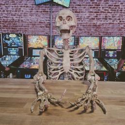A goofy lil' skeleton guy, sitting at a long table in the middle of Moby's. There is a row of old school pinball machines behind him. A goofy lil' skeleton guy, sitting at a long table in the middle of Moby's. There is a row of old school pinball machines behind him.