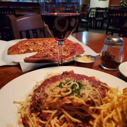 Spaghetti - The image shows a restaurant table with a glass of red wine, a slice of pizza, and a plate of spaghetti with meat sauce. The pizza is partially visible on a paper-lined tray, and the spaghetti is topped with cheese and herbs. The background includes empty chairs and tables, indicating a casual dining environment. The setting appears to be a sit-down restaurant with warm lighting.
