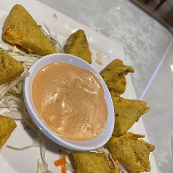 Bolly Twist - Appetizer - The image shows a white rectangular plate with several Indian samosas arranged around a small bowl of orange-colored dipping sauce. The samosas are golden-brown with a slightly greenish hue, indicating the use of spices or herbs in the dough. Shredded cabbage and carrots are placed underneath the samosas as garnish. The setting appears to be a casual dining environment, possibly an Indian restaurant, with a light-colored table surface. - Indian Near Me - Stuart, Florida
