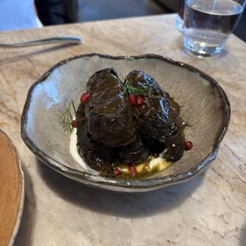 Rumis Kitchen - Dolmeh - A ceramic bowl contains dark, cooked vegetables garnished with herbs and pomegranate seeds, placed on a marble table in a restaurant setting. - Persian/Iranian Near Me - Houston, Texas