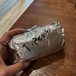 A person holds a foil-wrapped package with handwritten text 'T-Paco' on it, resting on a wooden table in a casual setting.