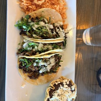 Cielo’s - Asada tacos - A rectangular white plate holds three tacos filled with seasoned meat, topped with chopped onions and cilantro. Accompanying the tacos is a portion of Mexican rice, and a chocolate dessert in a pastry shell with crumbled topping. The plate is placed on a wooden table, with a glass of water visible to the right. - Mexican Near Me - Snyder, Texas
