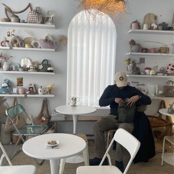Studio De Corner - Interior - A person wearing a beige cap, dark sweater, and beige pants sits at a small white table in a decorated interior space. The background features white shelves filled with ceramics, plush toys, and decorative objects. A large arched window with sheer curtains and a woven pendant light are also visible. The space has a cozy, minimalist aesthetic with neutral tones and natural textures. - Cafes Near Me - Markham, Ontario