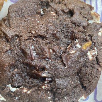 Insomnia Cookies - S'more's - A detailed view of a large chocolate cookie featuring chunks of chocolate embedded throughout. The cookie has a cracked surface with a slightly rough texture, indicating a baked, crispy exterior. It appears to be placed on a piece of parchment paper, with part of another baked item visible in the background. - Desserts Near Me - Albuquerque, New Mexico