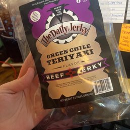 a hand holding a bag of beef jerky jerky jerky jerky jerky jerky jerky jerk - A hand holds a sealed plastic bag of beef jerky with a label from The Daily Jerky in Albuquerque, NM. The label indicates the flavor as Green Chile Teriyaki and includes branding, ingredients, and weight information. The background shows part of a keyboard and some notes.