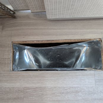 Shodeen - The image depicts a rectangular floor vent cover installed in a wooden floor. The vent cover has a reflective metal interior, which appears slightly wrinkled or bent. The surrounding flooring is light-colored wood, and part of a wicker basket and a textured wall are visible nearby. - Real Estate Services Near Me - Geneva, Illinois