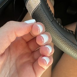 The image shows a person's left hand with a French manicure, featuring white tips and natural pinkish nails, holding a steering wheel inside a vehicle. The person is seated, with legs visible and wearing shorts. The steering wheel has a decorative band with a glittery or sparkly surface. The vehicle interior, including part of the dashboard and pedal, is also visible.