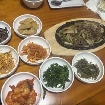 Sushi Box - Bulgogi - The image shows multiple small bowls of Korean side dishes, including kimchi, spinach, bean sprouts, and others, surrounding a sizzling plate of beef with onions. - Sushi Bars Near Me - Northbrook, Illinois