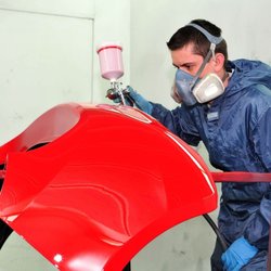 Gervic Auto Body Supply - Automotive painter in protective coveralls, gloves, and respirator sprays bright red paint onto a detached body panel mounted...