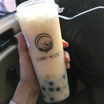 Starry Moon - Lemon Yakult - A person is holding a large plastic cup of bubble tea from Starry Moon, featuring a logo with a moon and a dragon. The drink appears to be a milk tea with black tapioca pearls at the bottom. The person is wearing a white wristband and is seated inside a vehicle, with the steering wheel and dashboard visible in the background. - Bubble Tea Near Me - Albuquerque, New Mexico