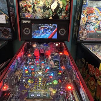 Atomic Age Modern - Pinball - A pinball machine themed around Elvira, House of Mystery, with colorful artwork depicting Elvira and spooky scenes. The machine has a digital display showing the game in progress, with illuminated bumpers, targets, and flippers. It is positioned among other arcade machines, indicating a recreational or arcade environment. - Thrift Stores Near Me - Mesa, Arizona