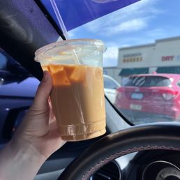 Iced white mocha with raspberry flavor and soy milk - A hand holds an iced coffee with milk and caramel in a clear plastic cup with a straw, inside a vehicle with a view of a shopping plaza outside.