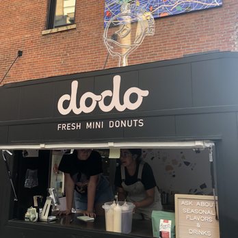 Dodo Donuts - the outside of a restaurant - A black food cart named 'dodo' specializes in fresh mini donuts, with a sign displaying the business name and offerings. Two staff members are visible inside the cart, preparing or taking orders. The cart features a sign encouraging customers to inquire about seasonal flavors and drinks. Above the cart, there is a colorful abstract artwork and a neon light sculpture of a hand holding a donut, attached to a brick building exterior. - Food Trucks Near Me - Columbus, Ohio