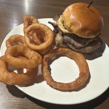 The Hideout Two - Brisket Burger and onion rings. This meal was amazing. - A burger with a bun, beef patty, and onions, served with onion rings on a white plate at a restaurant. - Seafood Near Me - Morris, Illinois