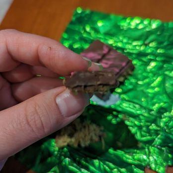 Sugar Bear - A bit blurry but chocolate on top was super thick, leaving not much room for the filling. - A hand with neatly trimmed nails holds a piece of chocolate bar, partially unwrapped from a shiny green foil wrapper. The background shows a wooden surface, and the foil wrapper is crinkled and reflective. The chocolate appears to be layered or filled, with some texture visible on the edges. - Candy Stores Near Me - Millburn, New Jersey