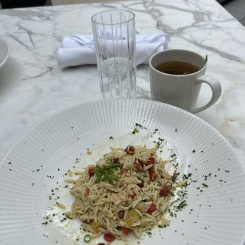 Flyleaf - Mediterranean Orzo Salad & Mint Tea - A plate of orzo pasta garnished with chopped herbs, vegetables, and toppings is placed on a marble table. Behind the plate, there is a glass of water, a cup of tea, and a napkin. The setting appears to be in a restaurant or cafe with a modern, elegant interior. - Bookstores Near Me - Grosse Pointe Farms, Michigan