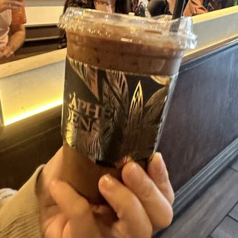 Caphe Den - A person is holding a plastic cup of iced coffee with a branded sleeve that features a design and text. The cup has a clear plastic lid and a straw. The background shows a cafe interior with a counter, a few people sitting and standing, and warm lighting. The setting appears casual and social. - Coffee & Tea Near Me - Flushing, New York