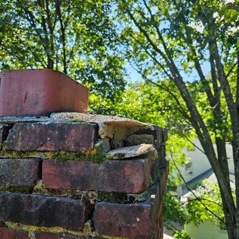 Tough Build Masonry - Before: Awful and falling apart. - The image features a close-up view of a brick chimney with moss growing between the bricks and a metal cap on top. The background includes green trees with sunlight filtering through the leaves and a partial view of a house. The scene suggests an outdoor setting during daytime. - Masonry/Concrete Near Me - Melrose, Massachusetts