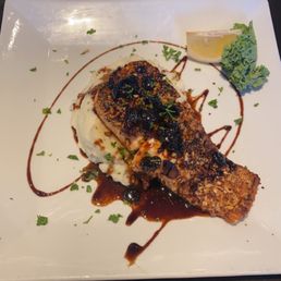 Almond crusted salmon - A dish with a piece of fish topped with sauce, served on mashed potatoes, garnished with herbs, lemon wedge, and a leaf of kale.