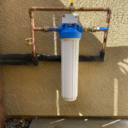 New Upgraded Nuvo Manor 2.0 system (with bypass and additional release valve) - A water filtration unit is mounted on an exterior wall, connected to copper and black pipes, with valves and a yellow handle on top.
