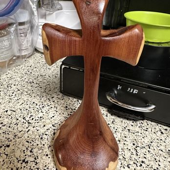 Buc-ee's - The beautiful soiled wooden cross  they sell in all different colors - A wooden cross sculpture with a polished finish and natural wood grain visible, standing upright on a speckled countertop. The sculpture has a simple, rustic design with rounded edges and a smooth surface, situated among kitchen items such as plastic bags, a blender, and a green bowl in the background. - Convenience Stores Near Me - Berthoud, Colorado
