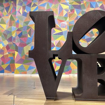 Newfields - Robert Indiana LOVE sculpture, gallery atrium (2nd floor) - A sculpture of the word 'LOVE' in large, black, three-dimensional letters is displayed in a museum with a colorful geometric mural behind it. - Parks Near Me - Indianapolis, Indiana