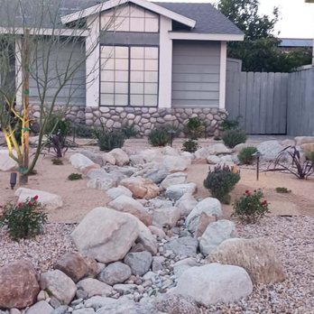 Smith Landscape - The image shows a landscaped front yard with a rock garden design, including large and small rocks, drought-tolerant plants, and a central tree. The yard is bordered by gravel pathways and low landscape lighting. A house with a stone foundation, siding, and a teal door is visible in the background, along with neighboring houses and a wooden fence. The scene emphasizes xeriscaping and low-maintenance landscaping. - Masonry/Concrete Near Me - Upland, California