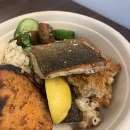 Market Bowl - A takeout meal in a disposable container includes a piece of grilled fish with crispy skin, macaroni and cheese, rice, cucumber slices, cherry tomatoes, and a lemon wedge. The food is arranged with the fish on top, surrounded by vegetables and sides, on a dark wooden surface.