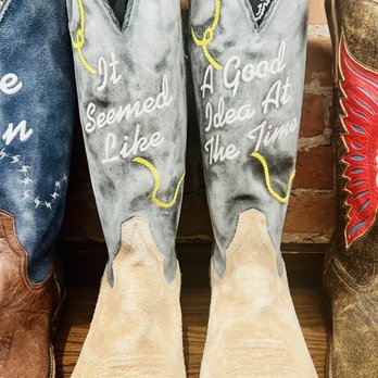 Kemo Sabe - always - Two cowboy boots with beige soles and gray upper sections are displayed side by side. The boots have handwritten phrases in white, with yellow stitching accents. The left boot reads, 'It seemed Like,' and the right boot says, 'A Good Idea At The Time.' The background includes a brick wall and other cowboy boots with different designs and colors. - Shoe Stores Near Me - Aspen, Colorado