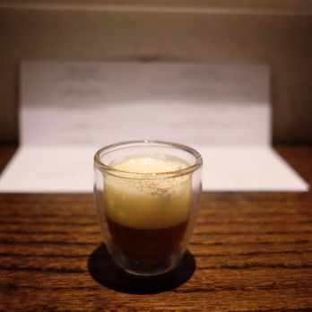Valhalla - French onion latte - A small glass cup holds a layered coffee drink with a dark coffee base and a frothy, creamy top layer. The drink is placed on a wooden table, with a blurred background that appears to be part of a menu or paper. The lighting highlights the drink and casts a shadow on the table. - Bars Near Me - Chicago, Illinois