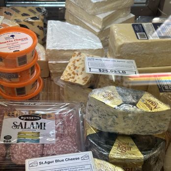 The Big Cheese - Big cheese: - The image shows a variety of cheeses, including blocks of white stilton with apricot, Saint Agur, and other cheese types, along with slices of cured salami. The cheeses are labeled with prices per pound, and some are wrapped in foil or plastic. The display is behind a glass window, with a wooden surface underneath, indicating a deli or cheese shop setting. - Cheese Shops Near Me - Annapolis, Maryland