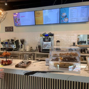Chai Corner - Zoom in for menus - A cafe counter features digital menu screens, coffee machines, and a display case with cookies and pastries, with a staff member visible through a window. - Breakfast & Brunch Near Me - Fairborn, Ohio