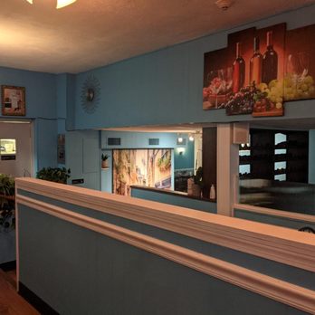 Ciao Amici Restaurant - Inside - The image shows the interior of a restaurant with blue-painted walls, decorated with artwork including a wine-themed painting and a scenic mural. Potted plants are placed on ledges and near the walls. The space includes a partial view of the dining area with tables and chairs, and warm lighting fixtures are visible. The overall decor is simple and inviting, with a focus on creating a cozy atmosphere. - Italian Near Me - Wakefield, Massachusetts