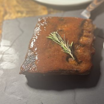 Le Chick - St. Louis BBQ Ribs - A cooked meat portion with a glossy glaze, garnished with a sprig of rosemary, resting on a dark, textured surface. The background includes a blurred bowl and a wooden surface, indicating a dining setting. - Cocktail Bars Near Me - Miami, Florida
