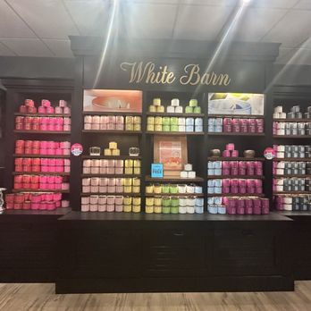 Bath & Body Works - A retail display at White Barn features multiple shelves filled with candles in different colors, including pink, yellow, green, blue, and gray. The candles are organized by color and size, with some promotional signs visible. The display is set against a dark background with the store's name, and the floor is wooden. The overall setting suggests a store dedicated to home fragrance products. - Cosmetics & Beauty Supply Near Me - Chicago, Illinois