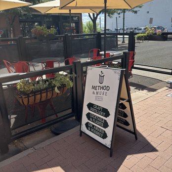 Method & Muse Spirits - Patio area with red chairs, tables, large umbrellas, and a sign advertising Method & Muse Spirits, craft distillery, cocktails, and whiskey. - Distilleries Near Me - Arvada, Colorado