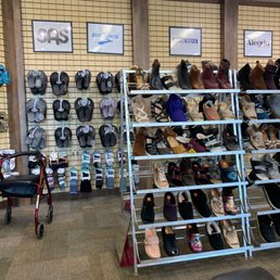 Inside - The image shows the interior of a shoe store with a display of women's footwear. Shoes such as sandals, boots, and various styles are arranged on a multi-tiered metal rack and wall hooks. The background features a wall with a grid pattern and four brand signs, including SAS, Brooks, Birkenstock, and Alegria. The store has a carpeted floor, a shopping cart, and a stool visible in the scene.