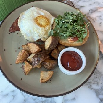 fl.2 - Eggs Benedict - A breakfast dish on a ceramic plate includes a fried egg with a runny yolk, roasted potato pieces, a sandwich topped with fresh greens, and a small cup of ketchup. The plate is placed on a marble table, with part of a person's green shirt visible in the background. - New American Near Me - Pittsburgh, Pennsylvania