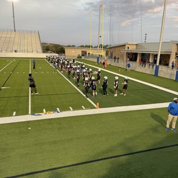 The Pfield - A group of football players in black and white uniforms are lined up on a football field, with some wearing helmets. Coaches and staff are present, and the field is marked with yard lines. The stadium has bleachers, lighting, and a building with spectators watching from the sidelines. The sky is partly cloudy, and the scene appears to be during late afternoon or early evening. - Stadiums & Arenas Near Me - Pflugerville, Texas