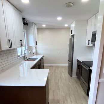 Adam Solutions - A kitchen with white upper cabinets and dark lower cabinets, stainless steel refrigerator and stove, and a light wood floor. The space includes a countertop with a sink, a window above the sink, and recessed ceiling lights. The layout is narrow with a clean, modern design. - Handyman Near Me - Macedon, New York