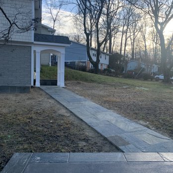 CM Hardscapes - Side stone walkway - A stone pathway extends through a yard, ending at a small white archway, with trees and residential houses in the background. - Masonry/Concrete Near Me