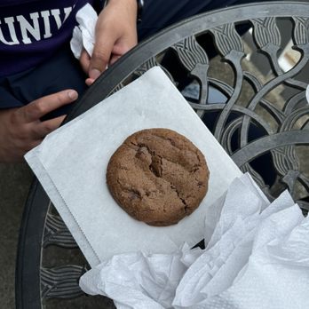 Coronation Cafe - Espresso chocolate chip cookie - A chocolate chip cookie is placed on a white napkin on a decorative metal outdoor table. The table has an intricate pattern, and part of a person's arm and hand are visible, resting nearby. The person is wearing a purple sweatshirt and a watch, and there are crumpled napkins on the table. - Coffee & Tea Near Me - Amherst, Massachusetts