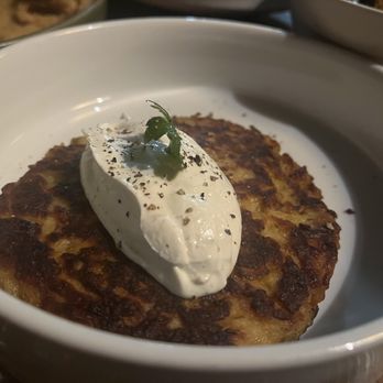 Sovana Bistro - Latkes - A golden-brown, crispy latke is presented in a white bowl, topped with a dollop of sour cream, black pepper, and a small green garnish. The latke appears freshly cooked with a textured surface, and the bowl is part of a dining setting with other dishes partially visible in the background. - Mediterranean Near Me - East Marlborough, Pennsylvania