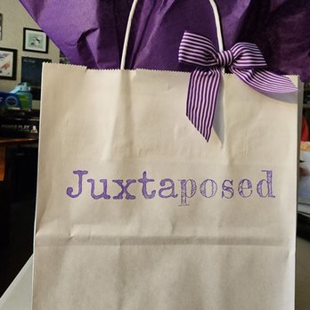 Juxtaposed - A beige paper gift bag features purple text spelling 'Juxtaposed' on its front. The bag has twisted paper handles, with a purple and white striped ribbon tied into a bow on one handle. Purple tissue paper is visible inside the bag, extending above the top edge. The background includes a room with framed pictures on the wall and various objects on a desk. - Home Decor Near Me - Albuquerque, New Mexico