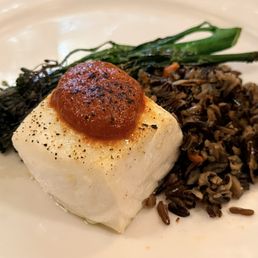 Halibut - The image shows a plated dish with a rectangular piece of white fish, topped with a roasted tomato. Accompanying the fish are green vegetables and a serving of dark rice, arranged on a white plate. The presentation emphasizes a balanced meal with seafood, vegetables, and grains.