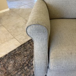 A beige fabric armrest of a sofa is shown, with a tiled floor and a patterned rug in the background.