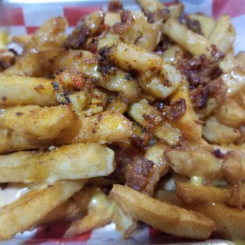 Neri's On the Square - Cajun Fries - A close-up image of French fries covered with melted cheese and sprinkled with bacon bits. The fries are golden and crispy, with cheese melted over them, and bits of bacon scattered on top. The food is served in a basket lined with red and white checkered paper, typical of casual dining or fast-food presentation. - New American Near Me - Graham, Texas