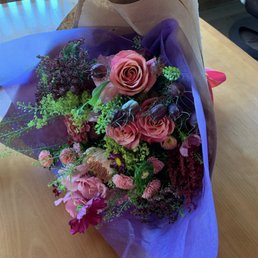 Hand tied bouquet florist choice - A floral bouquet featuring pink roses, carnations, and various greenery, wrapped in purple and brown paper, placed on a wooden table.